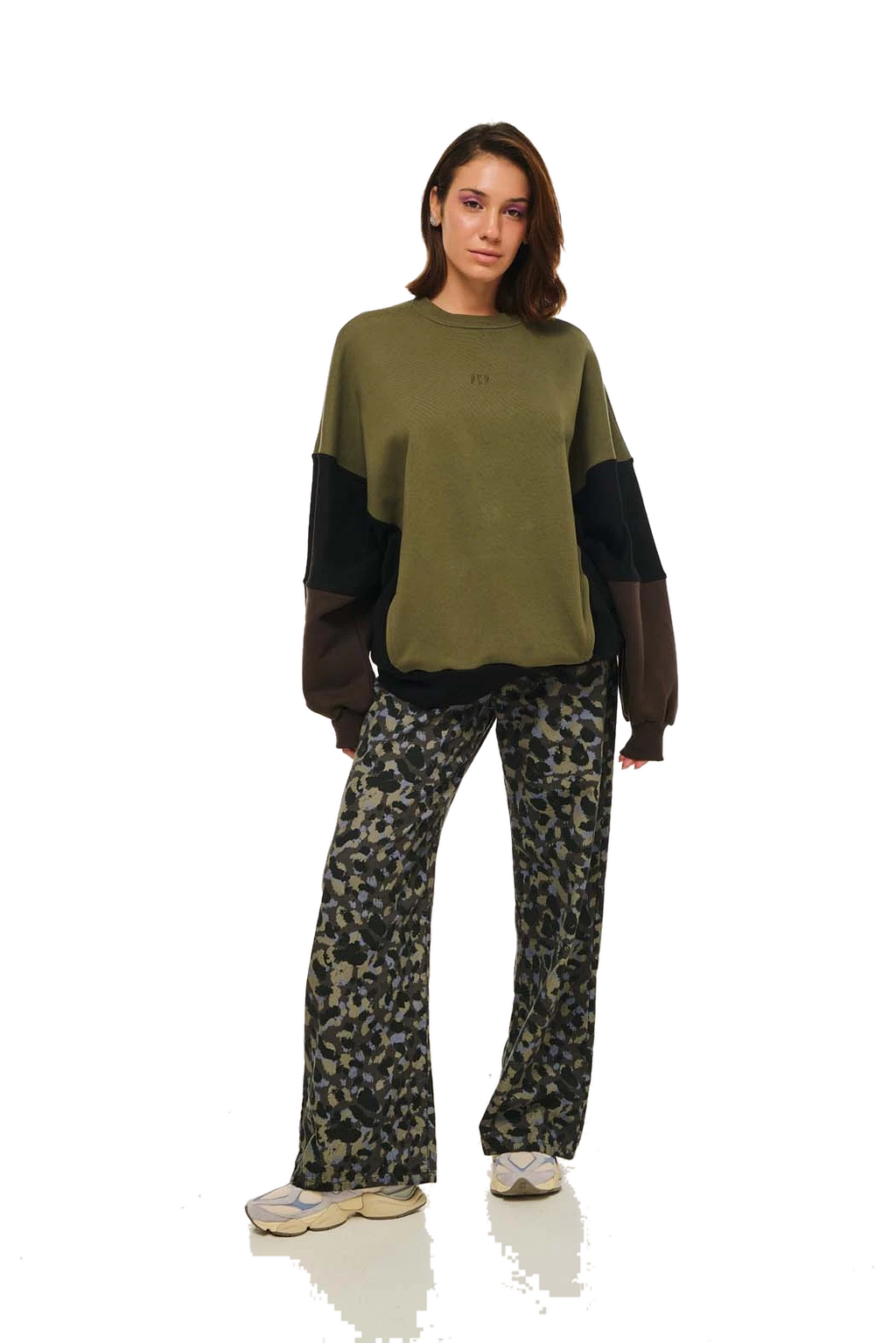 A model wearing a color-blocked crewneck sweatshirt in olive green and black, paired with dark camouflage print pants and beige sneakers, standing against a white background.