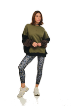 A woman modeling a color-blocked sweatshirt with olive green, dark brown, and black panels, paired with blue-gray patterned leggings and white sneakers, against a solid white background.