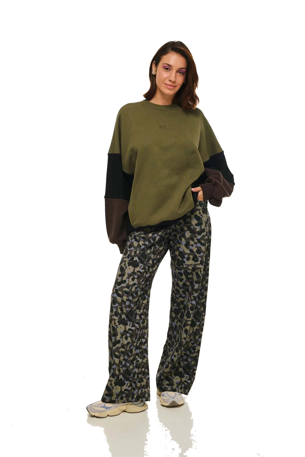 A woman modeling a color-blocked crewneck sweatshirt with olive green, black, and brown sleeves, paired with wide-leg camouflage pants and light-colored sneakers, against a white studio background.