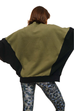 A person shown from the back wearing a color-blocked cape-style jacket with an olive green body and black sleeves, paired with blue-gray camouflage leggings, against a plain white background.