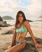 Woman in a green and white bikini sitting on a rock by the beach.
